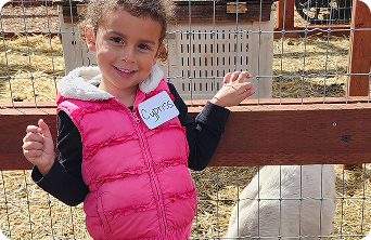 Young student wearing a pink vest smiling and standing by a fence during a farm field trip with a white goat in the background.