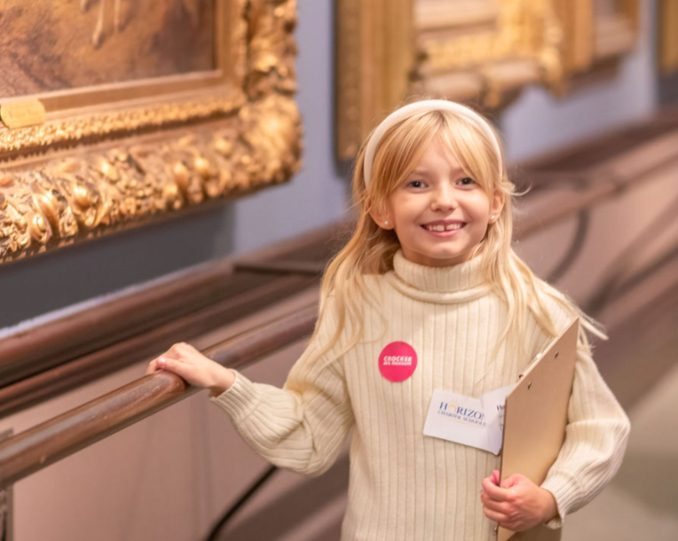 Young student smiling in an art museum while holding a clipboard, engaged in a learning activity with framed artwork displayed behind her.