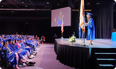 A student in a graduation gown speaking at a podium on stage, addressing an audience of seated graduates in caps and gowns during a ceremony.