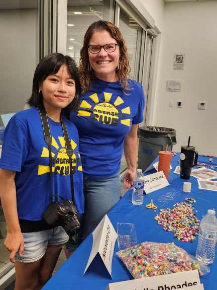 Female staff wearing leadership club tshirts with camera around their necks. Table in front of them has beads and name tags.