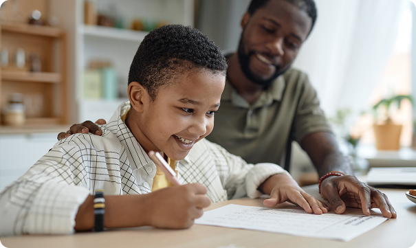 A smiling young student writes on a piece of paper at a desk while his parent watches supportively with a hand on the boy’s shoulder in a home setting.