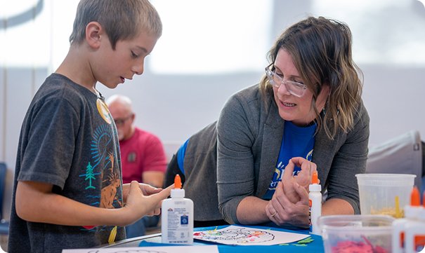 A female educator wearing glasses and a gray blazer leans in to assist a young boy with a craft project. The boy is looking down at his hands, and a bottle of white school glue sits on the table between them.