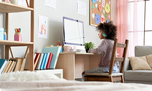 A young student wearing teal headphones sits at a desk in a bright room, focused on a large computer monitor while participating in remote learning.