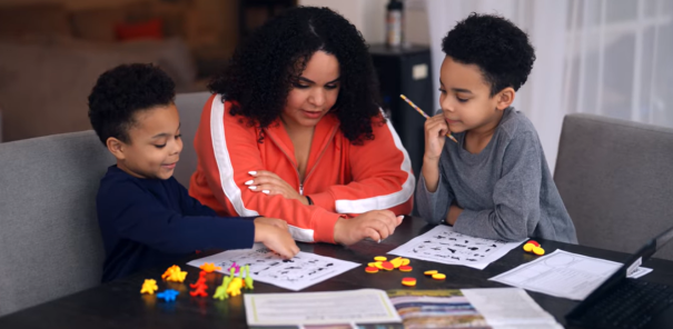 A mother sits at a table with her two young sons, helping them with educational worksheets and colorful learning manipulatives